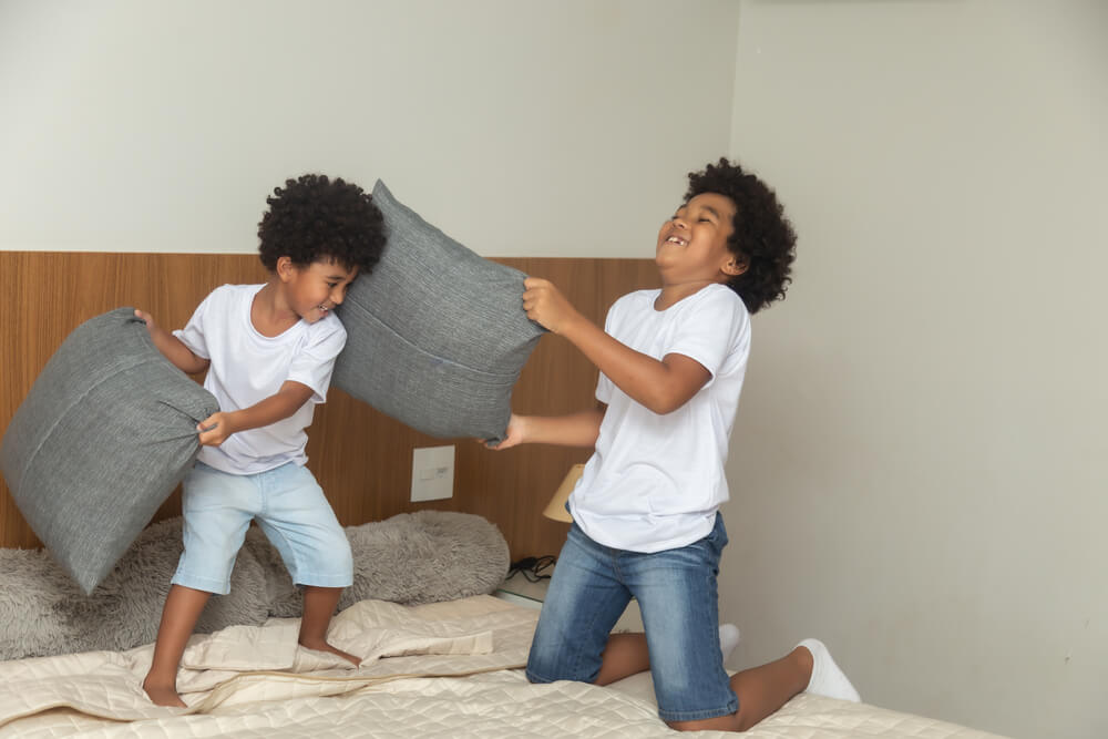 Duas crianças negras brincando de guerra de travesseiros em cima da cama. Elas estão gargalhando.