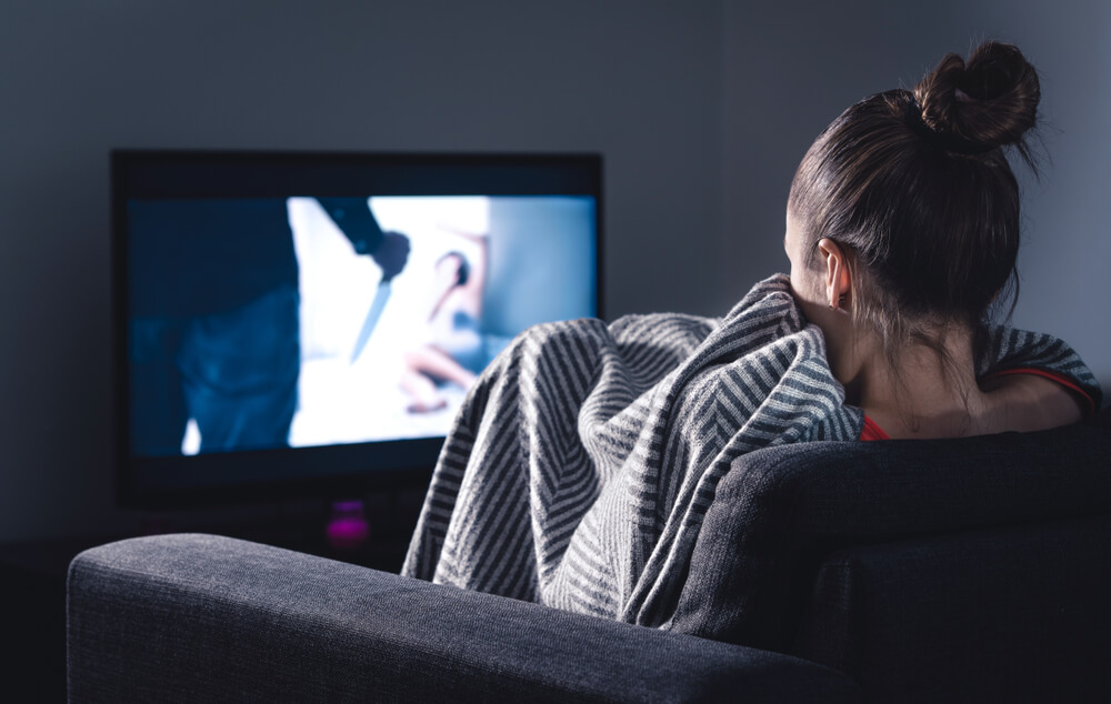 Imagem de uma mulher em frente a televisão de madrugada. Ela está enrolada em um cobertor. 