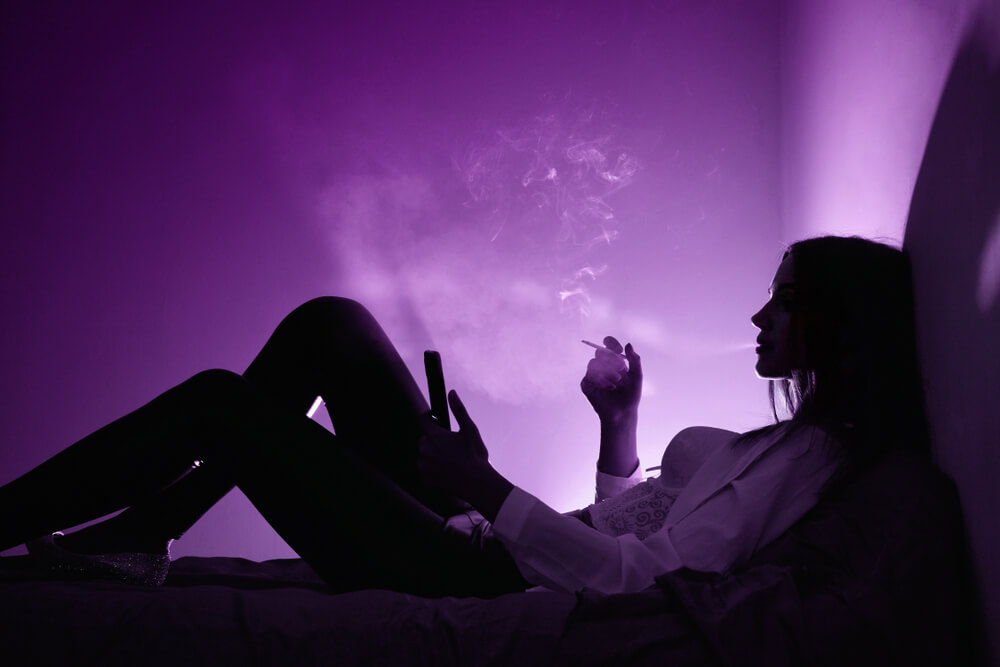 Imagem de uma mulher deitada na cama, fumando cigarro e mexendo no celular.