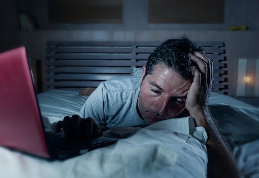 Imagem de um homem deitado na cama, aparentando sonolência, na frente de um computador.
