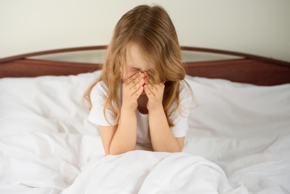 Menina sentada em uma cama, enquanto coça o nariz por causa de uma alergia.