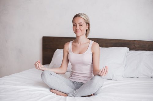 Mulher sentada em uma cama enquanto faz uma posição de Yoga e sorri.