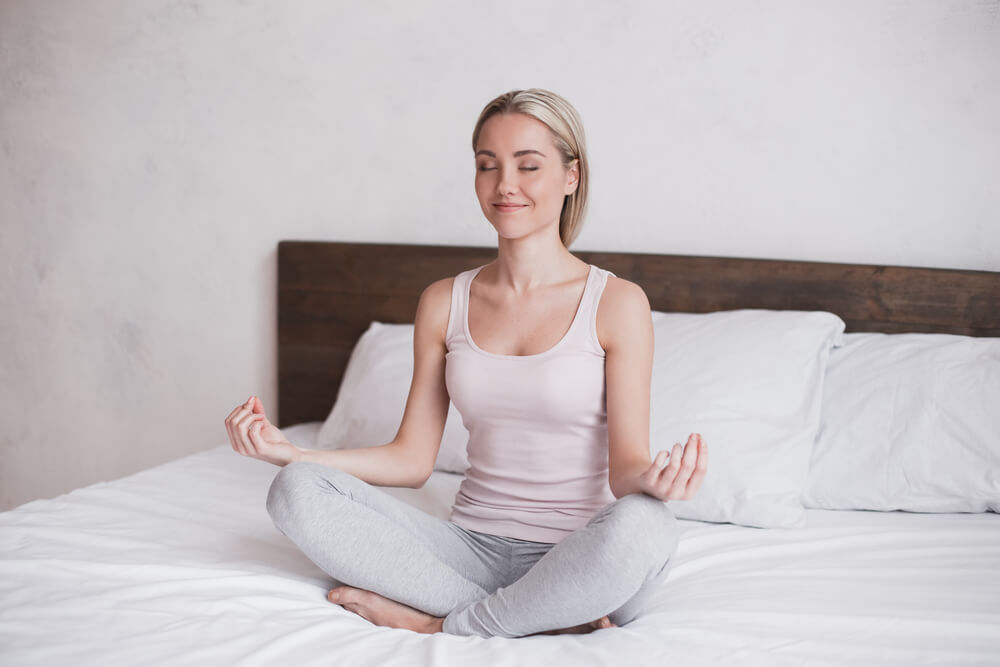 Mulher sentada em uma cama enquanto faz uma posição de Yoga e sorri.