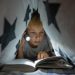 Criança deitada em uma cabana de lençol, segurando uma lanterna que está iluminando um livro.