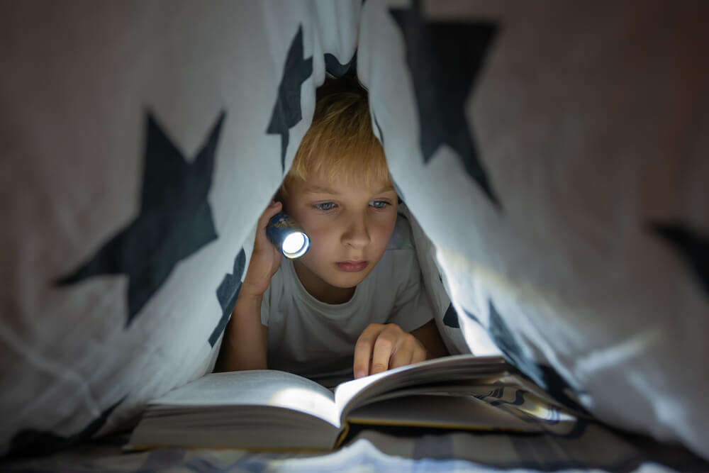 Criança deitada em uma cabana de lençol, segurando uma lanterna que está iluminando um livro.