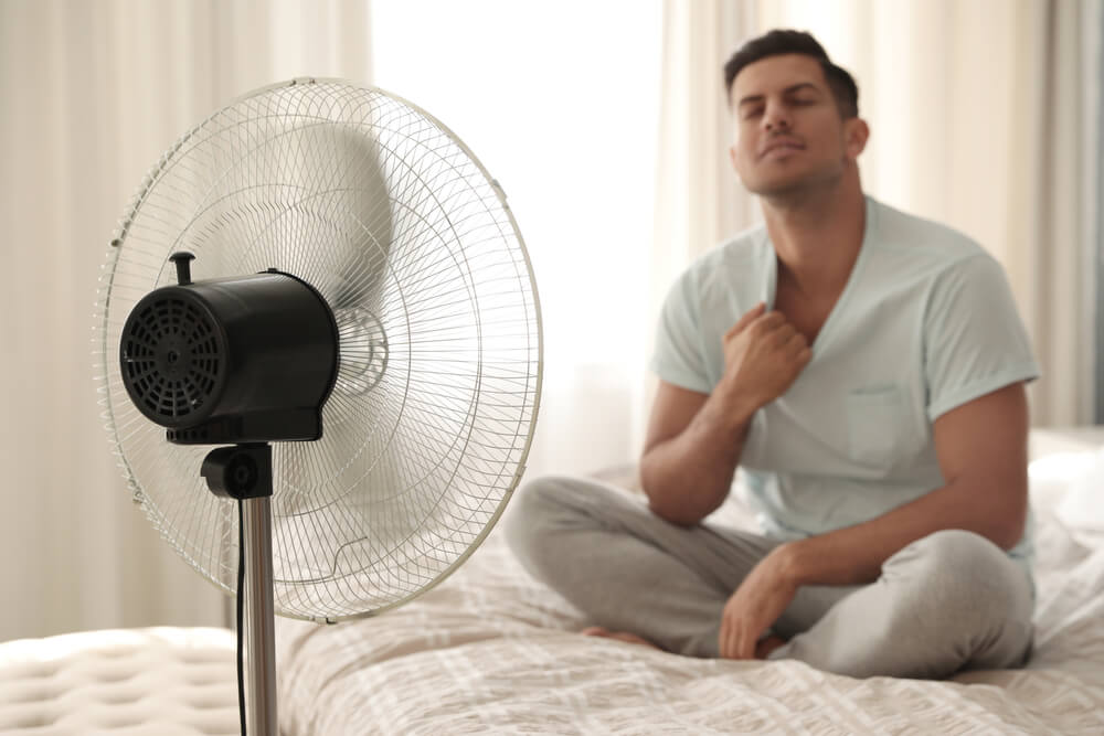 um homem sentado em uma cama ao lado de um ventilador
