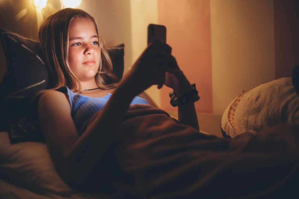 Imagem de uma garota adolescente. Ela está deitada na cama e utilizando o celular.