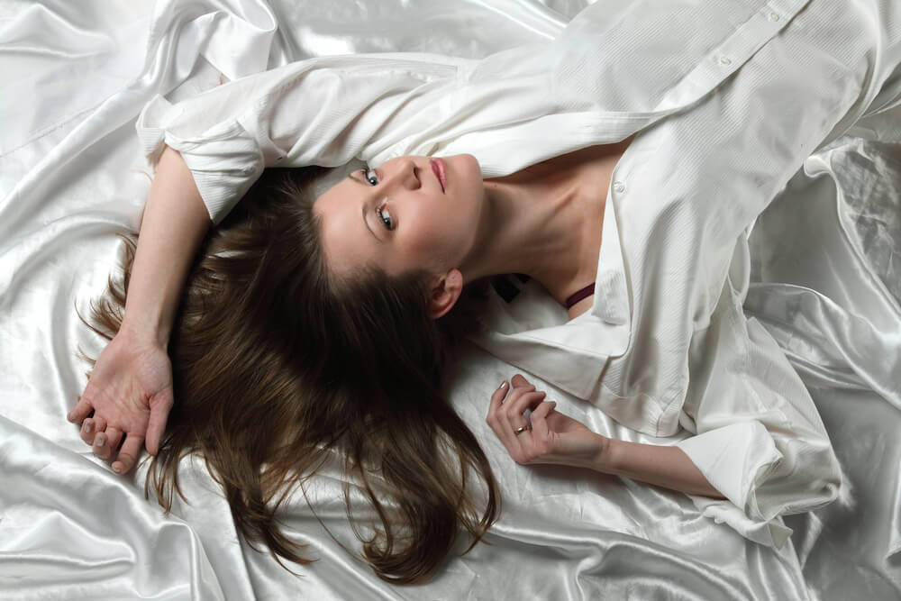 Imagem de uma mulher deitada na cama, com os cabelos soltos, em um lençol de cetim.