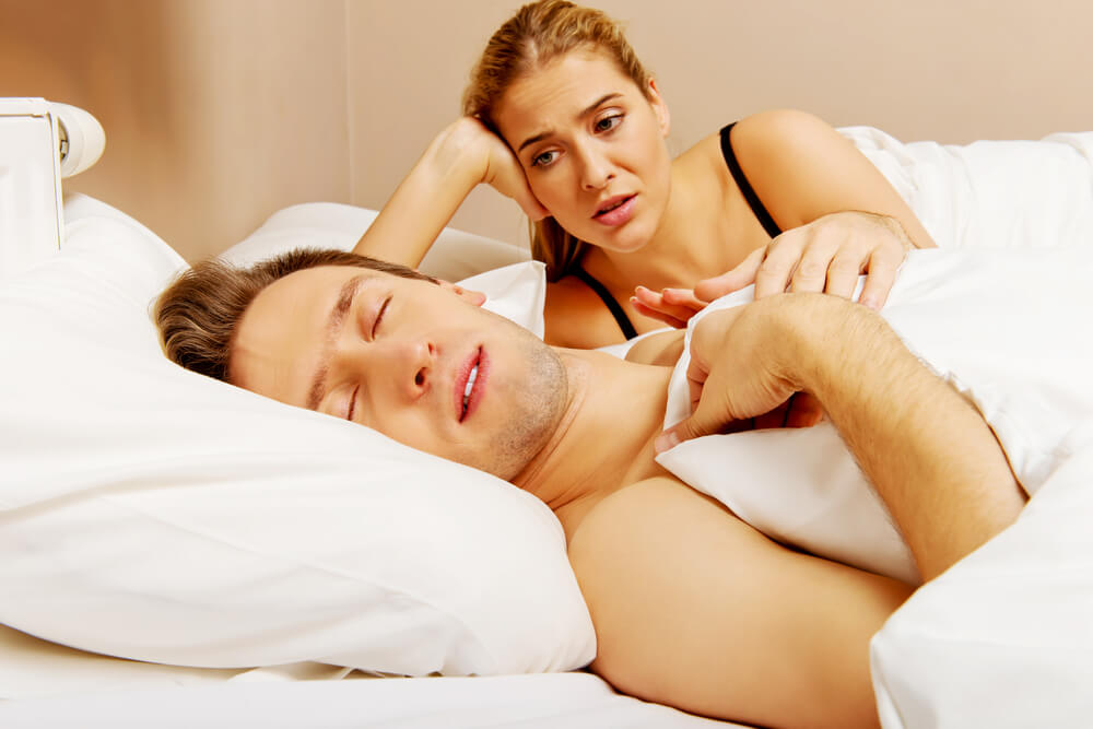 Homem deitado na cama, com a boca aberta, enquanto uma mulher ao lado dele está acordada e mexendo nele.