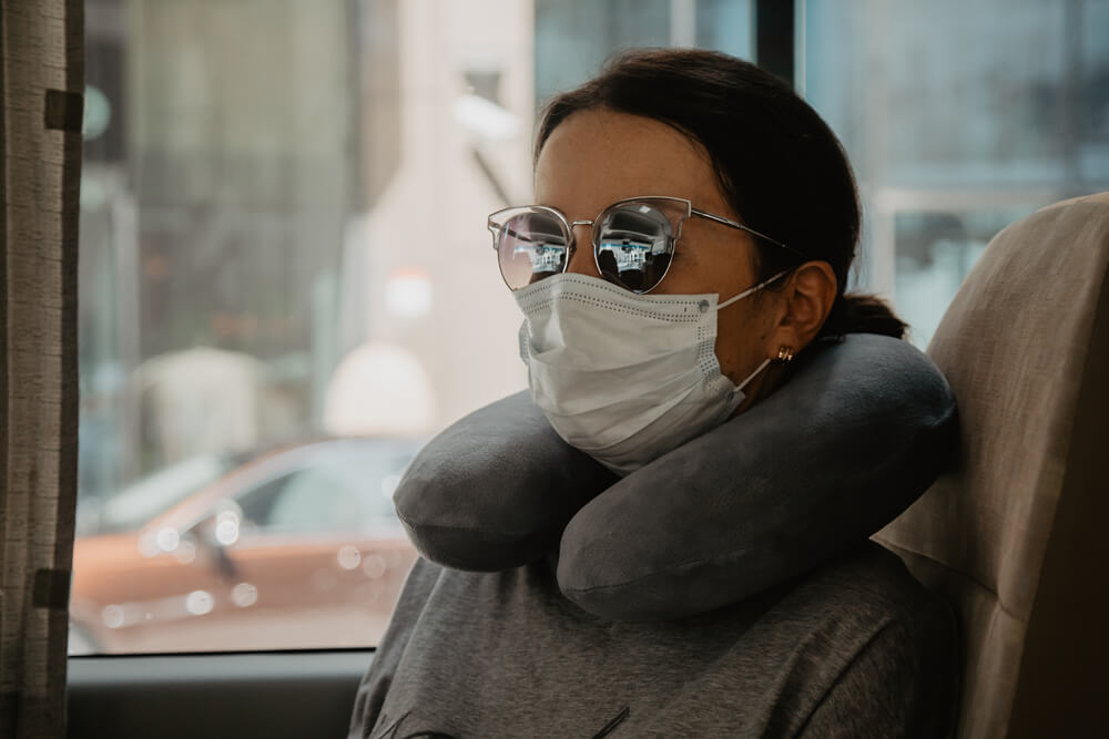 Mulher dentro de um ônibus. Ela está de máscara, com um óculos e um travesseiro de pescoço, pronta para dormir no ônibus.