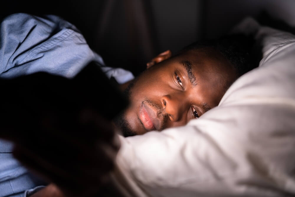 Homem negro, deitado na cama, mexendo no celular.