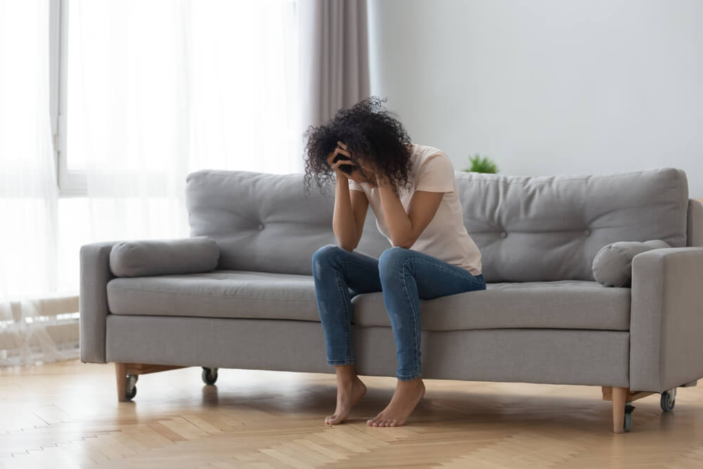 Imagem de uma mulher negra. Ela está sentada em um sofá e com as mãos na cabeça, demonstrando cansaço e irritabilidade. 