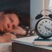 A imagem mostra uma pessoa deitada na cama, acordada, olhando o relógio, sem conseguir dormir.