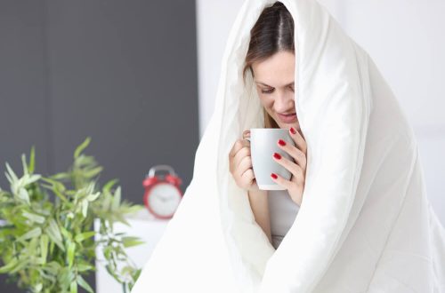 Imagem de uma mulher embaixo de uma coberta, com uma xícara de chá nas mãos, pronta para dormir no frio.