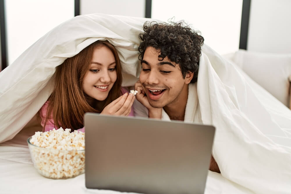 Imagem de um garoto e uma garota. Eles estão deitados na cama, em frente a um notebook, comendo pipoca e assistindo a filmes de inverno.