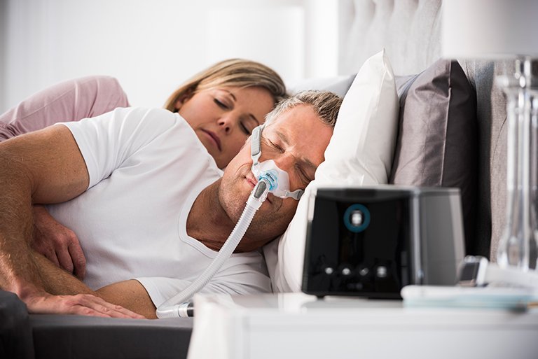 um homem e uma mulher deitados na cama com um tubo de oxigênio preso ao lado